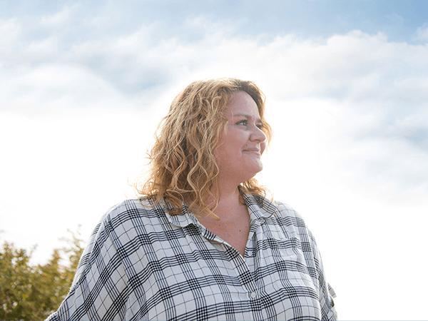 Stock photo of a plus-sized white woman with curly blonde hair smiling in front of a bright but cloudy sky as she looks into the distance