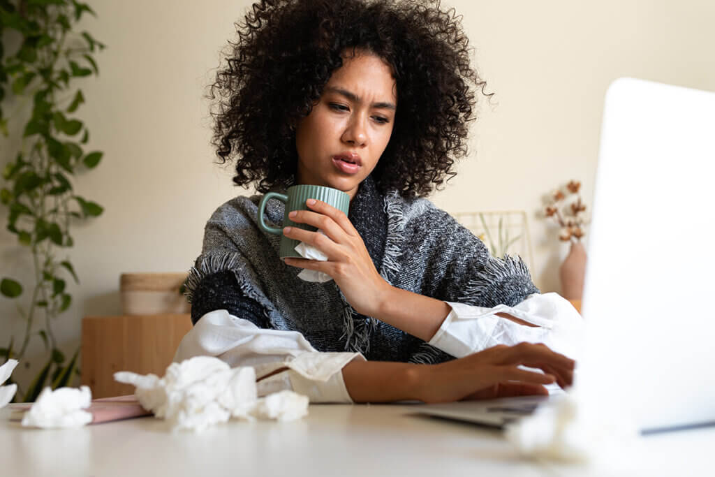 Sick African American woman working from home office using laptop drinking hot beverage covered with blanket.